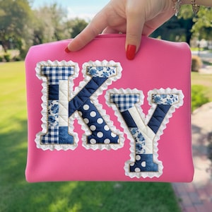 Puede incluir: Una bolsa rosa con las letras "KY" aplicadas. Las letras están hechas de varias telas azules, incluyendo lunares, estampados florales y cuadros de vichy, y están bordeadas con encaje blanco.