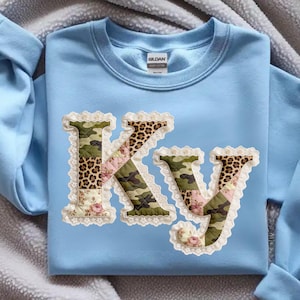 Può includere: Felpa azzurra con le lettere "KY" applicate sul davanti. Le lettere sono in stile patchwork, con motivi mimetici, leopardati e floreali, rifinite con pizzo bianco.