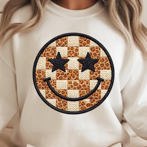 Op de afbeelding: Crèmekleurige sweatshirt met een groot, geborduurd smiley-gezicht. De smiley heeft zwarte sterogen, een zwart gestikt lachje en een geblokt patroon met luipaardprint accenten. Het ontwerp is omlijst door een zwarte rand.
