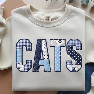 May include: Cream-colored sweatshirt with the word "CATS" appliqued in a patchwork design. The letters feature various patterns, including gingham, polka dots, and floral prints, with paw prints and a bow. The sweatshirt has a crew neck and a Comfort Colors tag.