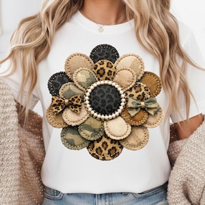 Può includere: T-shirt bianca con un grande motivo floreale. Il fiore è composto da varie toppe di tessuto nei toni del marrone, beige, nero e mimetico, con un centro perlato e due piccoli fiocchi. Il disegno è cucito sulla maglietta.