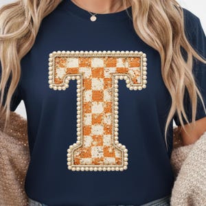 May include: Navy blue t-shirt featuring a large, decorative letter "T". The letter is filled with an orange and white checkered pattern and outlined with small, pearl-like beads. The shirt has short sleeves.