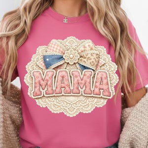 Peut inclure: T-shirt rose avec un motif décoratif. Le motif présente le mot "MAMA" en lettres roses avec une bordure crème. Le motif comprend un cadre ovale en dentelle et un nœud avec des motifs vichy, denim et floral.