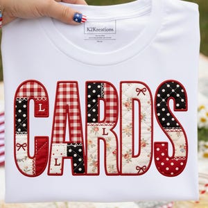 May include: White t-shirt with the word "CARDS" in a patchwork design. The letters are outlined in red and feature patterns like gingham, polka dots, and floral prints. The shirt is held up by a hand with a patriotic nail design.