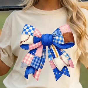 Puede incluir: Camiseta color crema con un gran diseño de lazo colorido. El lazo es un patchwork de telas azules, rosas y marrones, con costuras inspiradas en el béisbol y un centro azul. El lazo es el punto focal de la camiseta.