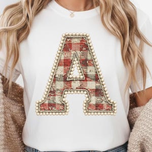 Könnte beinhalten: Weißes T-Shirt mit einem großen, dekorativen Buchstaben "A". Der Buchstabe hat ein rot-cremefarbenes Karomuster, das mit Kunstperlen und einer seilartigen Umrandung versehen ist. Ein lässiger, alltagstauglicher Stil.