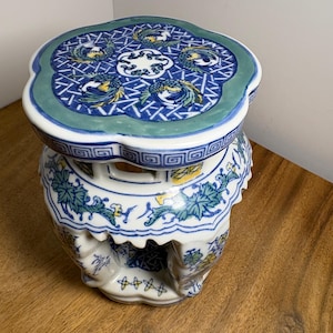 May include: A ceramic garden stool featuring a blue, green, and yellow floral and bird pattern. The top surface has a scalloped edge with a central blue and white design, while the sides display cut-out sections and intricate painted details.