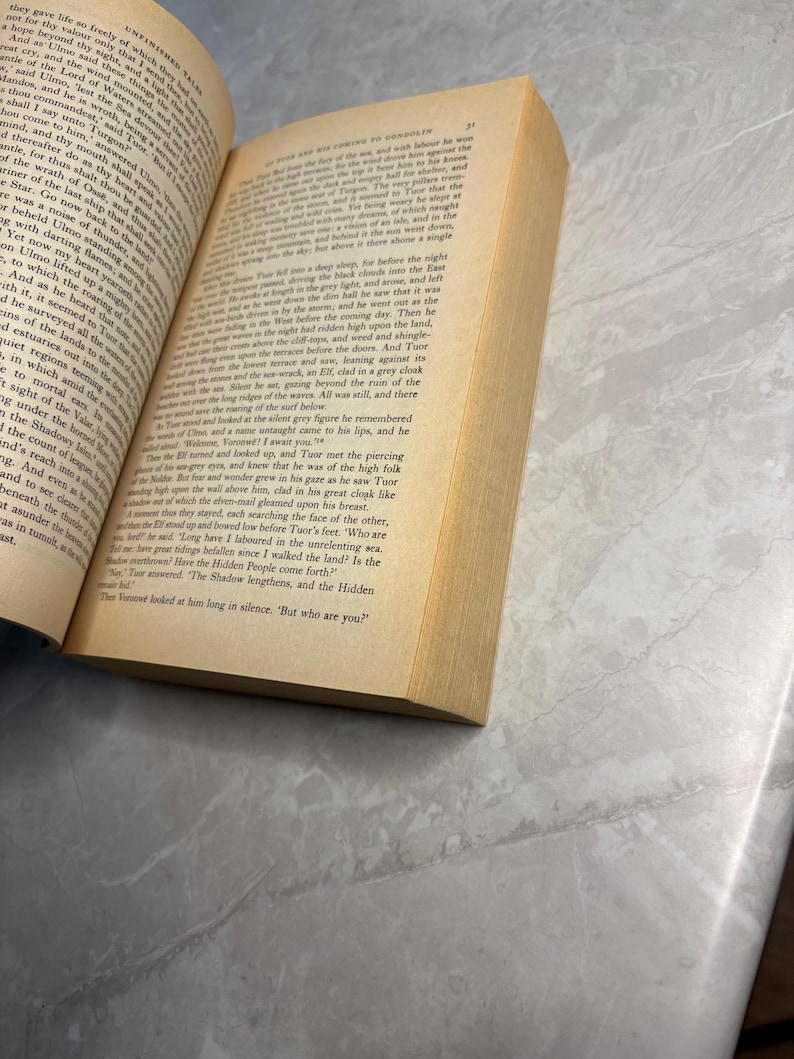 May include: An open book with yellowed pages, titled "Unfinished Tales." The text is in English, with a focus on the story. The book is on a light gray surface.