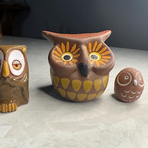 May include: Three ceramic owl figurines of varying sizes. The left owl is brown with large white eyes. The centre owl is brown with orange-rimmed eyes and yellow chest markings. The right owl is a small, round brown figure with white eye details.