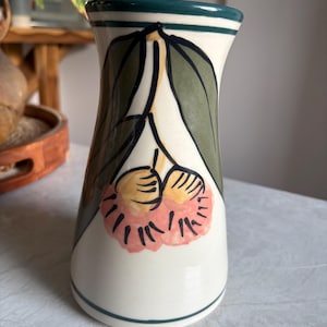 May include: A hand-painted ceramic vase with a white base, featuring a floral design with green leaves and pink and yellow flowers. The vase has a dark green rim and base trim, and is suitable for home decor.