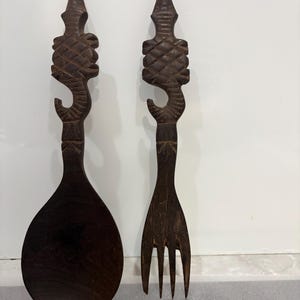 May include: A dark brown wooden spoon and fork set. The handles have carved, ornate designs with a hook shape. The spoon has a large, rounded bowl, and the fork has four tines. The set is likely for serving food.