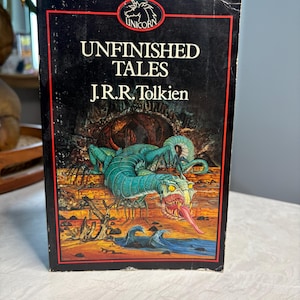 May include: A vintage paperback book titled "Unfinished Tales" by J.R.R. Tolkien. The cover features a fantasy illustration of a serpentine creature with a long red tongue, set against a backdrop of a landscape with water and a red border.