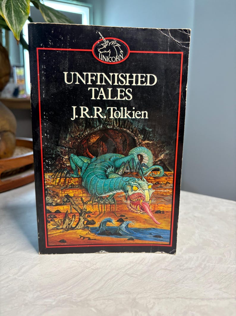 May include: A vintage paperback book titled "Unfinished Tales" by J.R.R. Tolkien. The cover features a fantasy illustration of a serpentine creature with a long red tongue, set against a backdrop of a landscape with water and a red border.