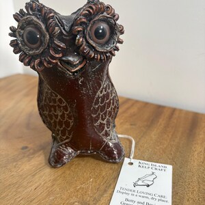 May include: A dark brown owl figurine with large, round eyes and textured feather details. A white rectangular tag is attached, reading: KING ISLAND KELP CRAFT, TENDER LOVING CARE: Display in a warm, dry place. Betty and Bevin Grassy, King Island (03) 6461 1464.