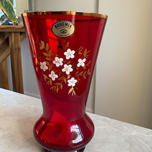 May include: A ruby red Bohemian glass vase with a gold rim and a flared shape. The vase features a floral design with white flowers and gold leaves. The label reads "Bohemia Czechoslovakia Hand Made."