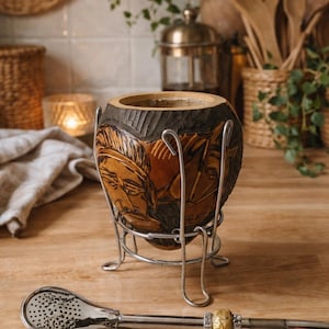 Peut inclure: Une calebasse artisanale avec un motif sculpté de visages et de chevaux, maintenue dans un support en métal argenté. Une bombilla argentée avec un accent doré décoratif repose sur une surface en bois. La calebasse est pour boire du yerba maté.