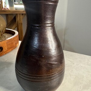 May include: A dark brown ceramic vase with a wide base and a narrow neck. The vase has horizontal grooves around the body and neck. The surface has a glossy finish.