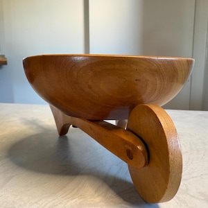 May include: A wooden bowl in a wheelbarrow design. The bowl is made of light brown wood and has a rounded shape. The wheel is also made of wood and is attached to the bowl with a wooden support.