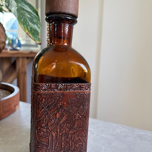 May include: Amber glass bottle with a wooden stopper and a brown leather sleeve. The leather sleeve features a detailed embossed scene of figures on horseback. A small gold chain connects the stopper to the bottle.