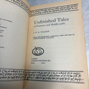 May include: Open book titled "Unfinished Tales of N&uacute;menor and Middle-earth" by J.R.R. Tolkien. The book's pages are off-white with black text and decorative borders. The publisher is Unwin Paperbacks, with a unicorn logo.