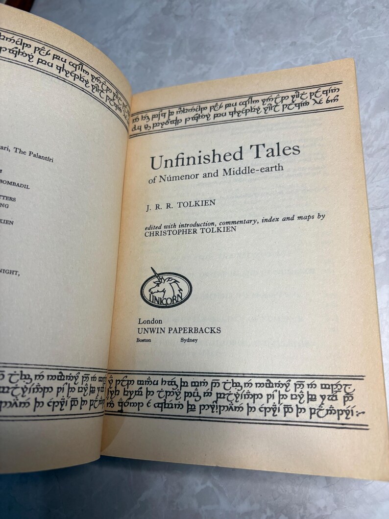 May include: Open book titled "Unfinished Tales of N&uacute;menor and Middle-earth" by J.R.R. Tolkien. The book's pages are off-white with black text and decorative borders. The publisher is Unwin Paperbacks, with a unicorn logo.