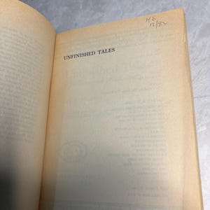 May include: Open book with the title "UNFINISHED TALES" printed on the page. The book's pages are aged with a yellowed hue. Handwritten text "HS 12/82" is visible in the upper right corner.