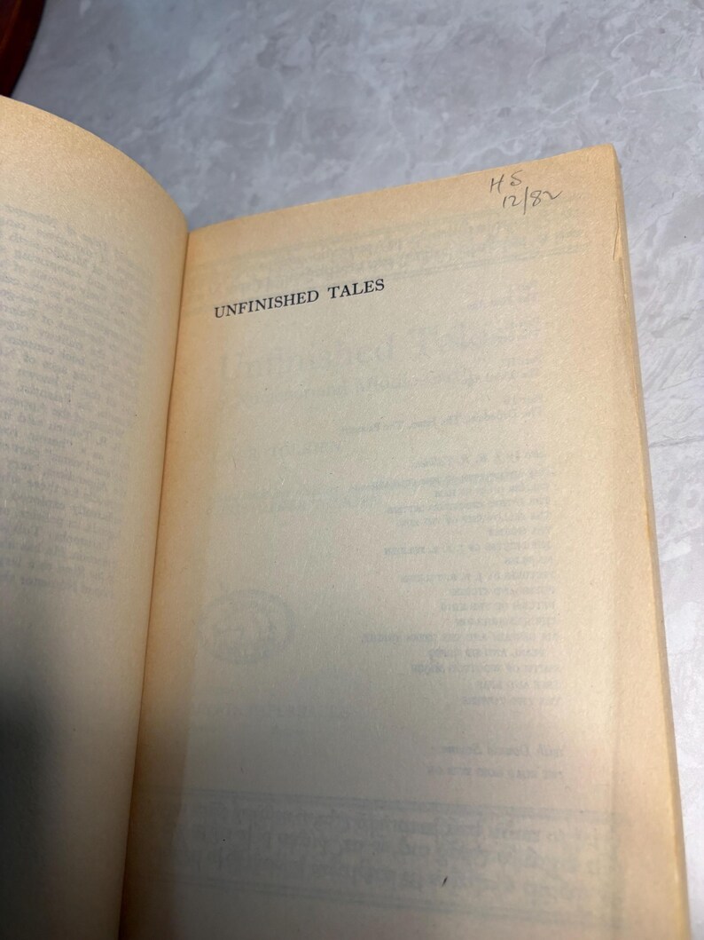 May include: Open book with the title "UNFINISHED TALES" printed on the page. The book's pages are aged with a yellowed hue. Handwritten text "HS 12/82" is visible in the upper right corner.