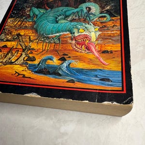 May include: A vintage hardcover book titled "Tales" by J.R.R. Tolkien. The cover features a colorful illustration of a turquoise dragon-like creature with a long pink tongue, set against a landscape with waves and a rocky shore.