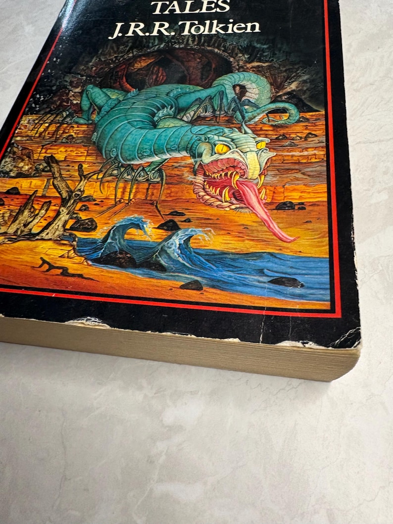 May include: A vintage hardcover book titled "Tales" by J.R.R. Tolkien. The cover features a colorful illustration of a turquoise dragon-like creature with a long pink tongue, set against a landscape with waves and a rocky shore.