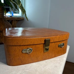 Pode incluir: Uma mala vintage castanha retangular com bordas arredondadas. A mala tem um acabamento brilhante e é presa com um fecho cor de latão e duas travas laterais. A mala é provavelmente de meados do século XX.