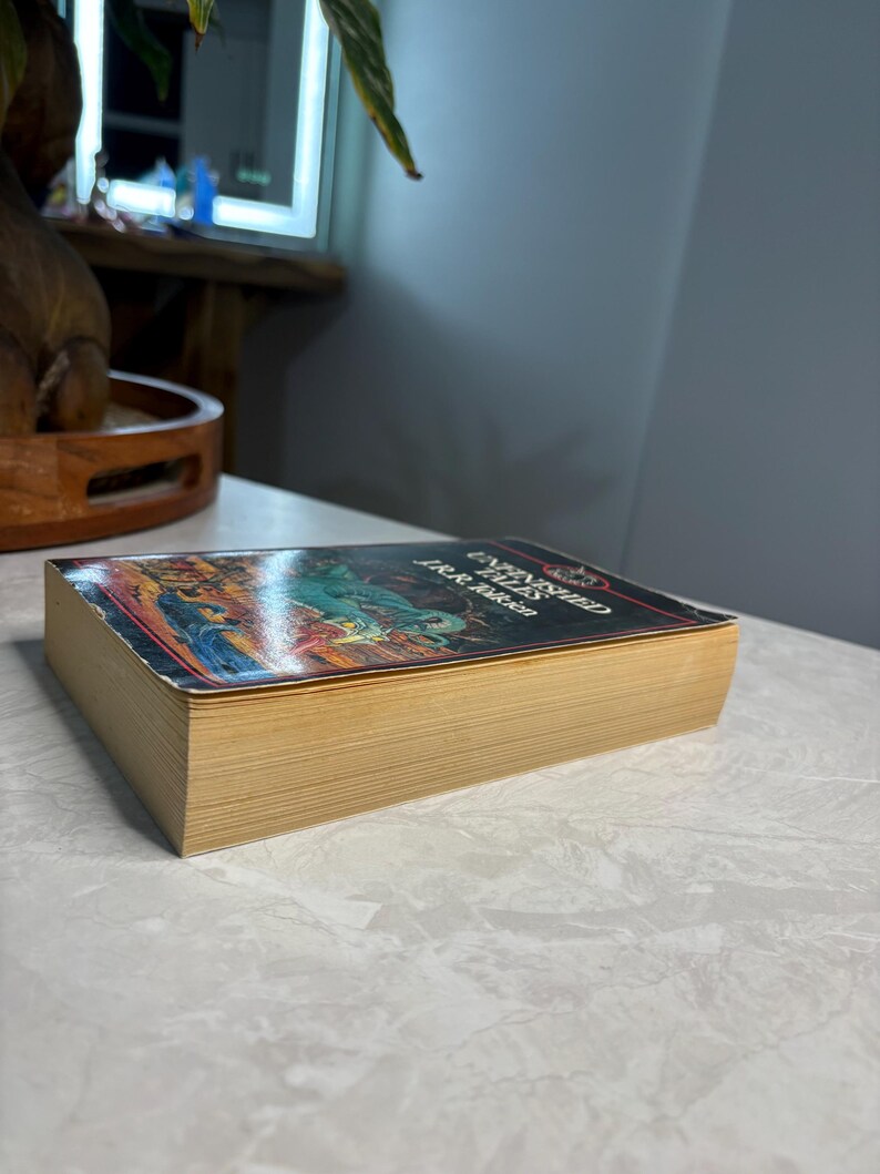 May include: A hardcover book titled "Unfinished Tales" by J.R.R. Tolkien. The book has a colorful cover with illustrations and gold-colored page edges. The book is lying on a white surface.