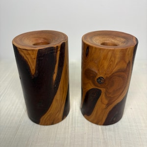 May include: Two wooden candle holders with a natural, organic design. The candle holders are cylindrical with a dark brown and light brown wood grain pattern. Each has a circular indentation at the top for a candle.