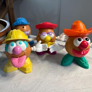 May include: Four Mr. Potato Head toys with different outfits and accessories. One wears a yellow construction hat and outfit, another a red hat, and an orange cowboy hat. Each has white hands and various facial features.