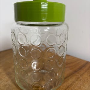 May include: A clear glass storage jar featuring a repeating embossed circular pattern across its exterior surface. The jar is topped with a solid, bright lime green lid that includes a small, rounded knob handle at the very centre of the top.