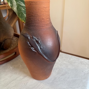 May include: A terracotta vase with a textured neck and a dark brown body. A detailed lizard design wraps around the vase. The vase is a decorative item.