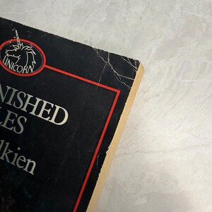May include: A close-up of a book titled "Unfinished Tales" by J.R.R. Tolkien. The book has a black cover with white text and a red border. The Unicorn logo is visible. The book's edges are worn.