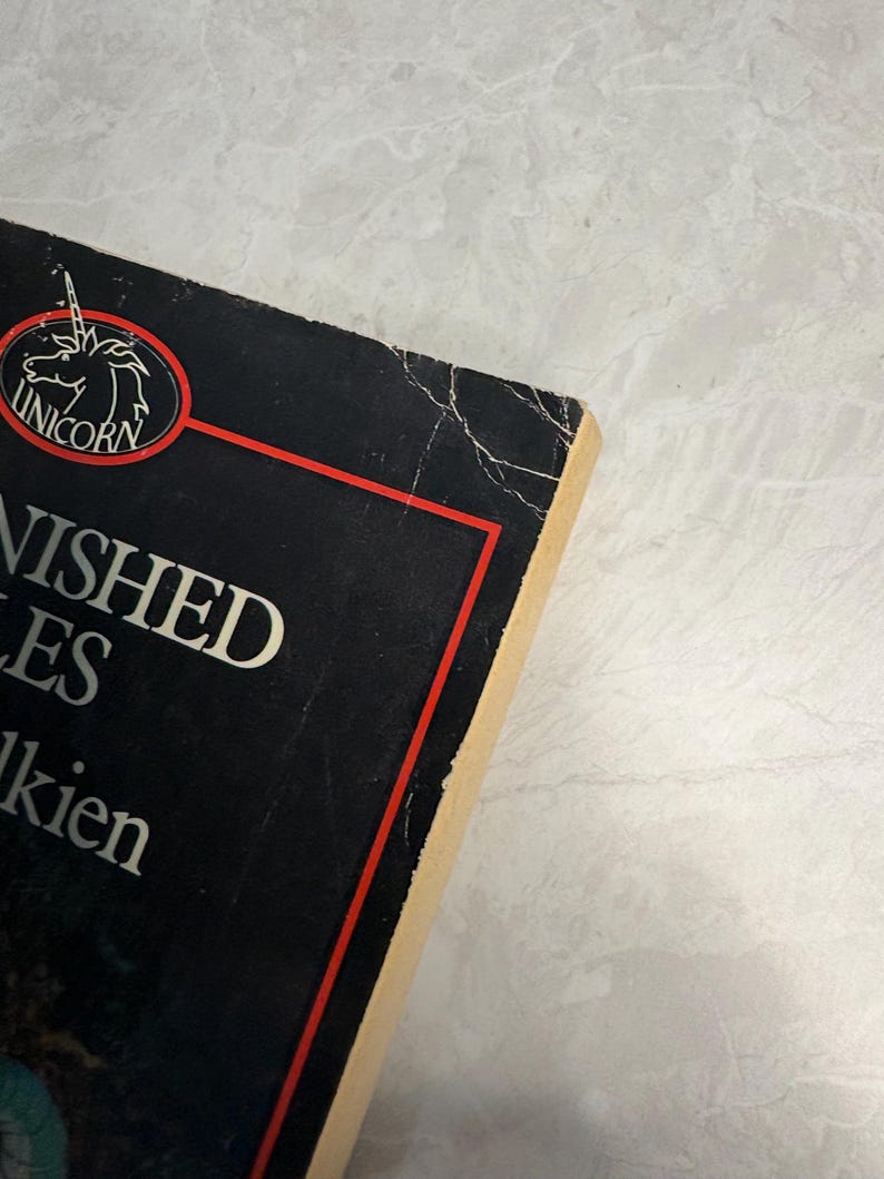 May include: A close-up of a book titled "Unfinished Tales" by J.R.R. Tolkien. The book has a black cover with white text and a red border. The Unicorn logo is visible. The book's edges are worn.