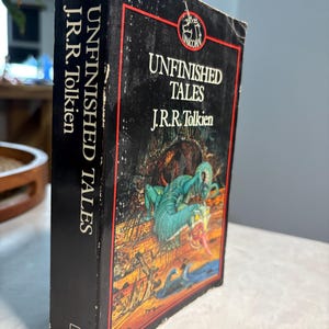 May include: A hardcover book titled "Unfinished Tales" by J.R.R. Tolkien. The book has a black cover with a red border and a fantasy illustration of a dragon. The spine displays the title and author's name in white.