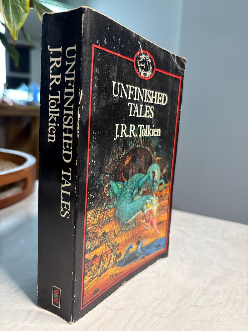 May include: A hardcover book titled "Unfinished Tales" by J.R.R. Tolkien. The book has a black cover with a red border and a fantasy illustration of a dragon. The spine displays the title and author's name in white.