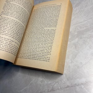 May include: An open book with yellowed pages, featuring printed text. The book's spine is visible, and the pages are slightly curved. The book is resting on a light-colored surface.