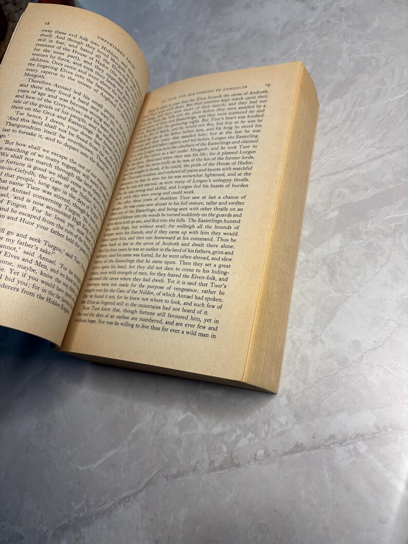 May include: An open book with yellowed pages, featuring printed text. The book's spine is visible, and the pages are slightly curved. The book is resting on a light-colored surface.