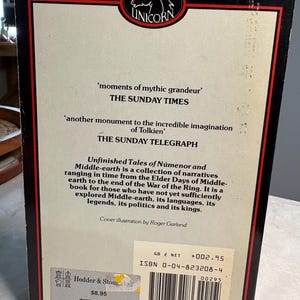 May include: The back cover of a book titled "Unfinished Tales of N&uacute;menor and Middle-earth." The cover features quotes from The Sunday Times and The Sunday Telegraph. The book has a black border with a red inner frame and a Unicorn logo.