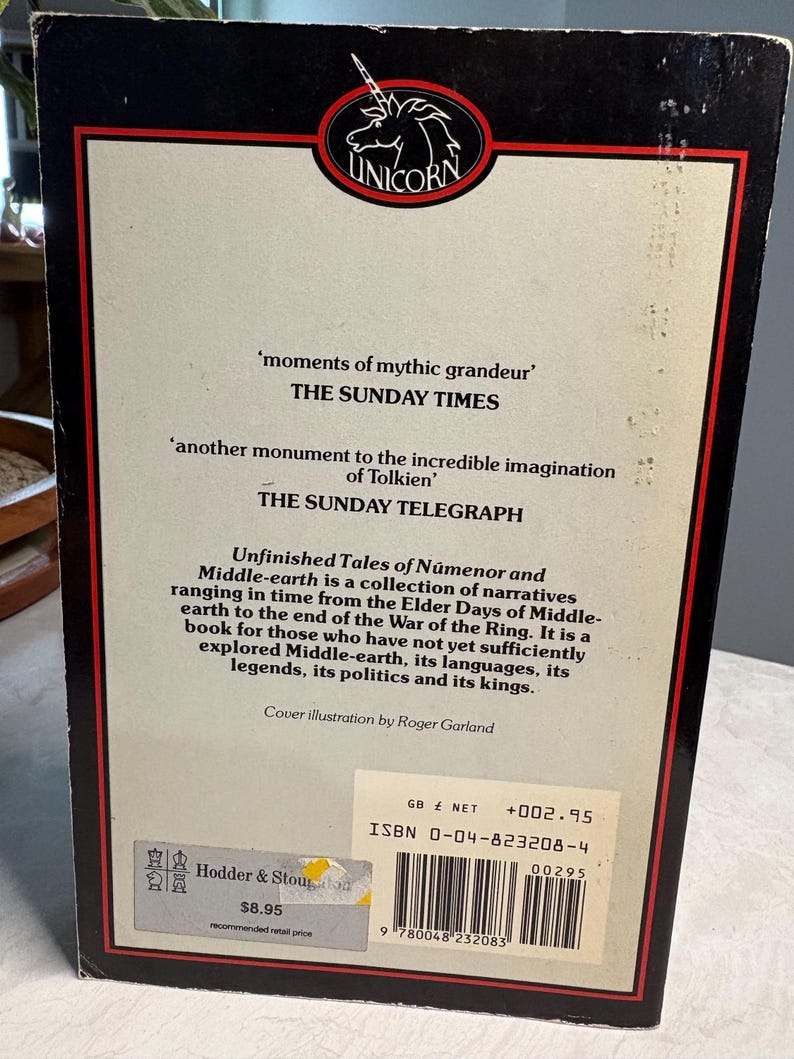 May include: The back cover of a book titled "Unfinished Tales of N&uacute;menor and Middle-earth." The cover features quotes from The Sunday Times and The Sunday Telegraph. The book has a black border with a red inner frame and a Unicorn logo.