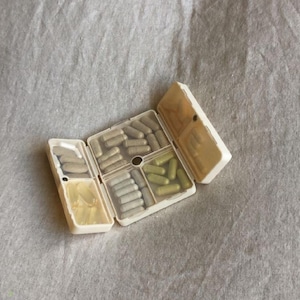 Puede incluir: Un pastillero de plástico beige, abierto para revelar varias cápsulas y cápsulas blandas. El organizador está dividido en compartimentos, algunos con cápsulas de color marrón claro, otros con cápsulas verdes y cápsulas blandas transparentes. El fondo es una tela gris claro.