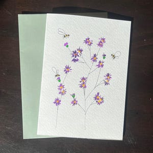 May include: A handmade greeting card featuring a watercolor illustration of purple flowers and bees. The card is paired with a light green envelope. The artwork is on textured white paper, with a dark brown background.