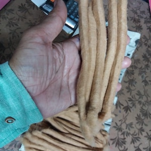 May include: A hand holds several bundles of light brown, fibrous material, possibly cotton or wool. The material is arranged in long, thick strands. A calculator and part of a teal shirt are visible in the background.