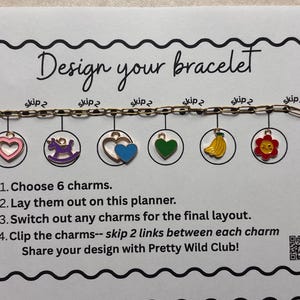 May include: A gold-toned metal chain bracelet with six colorful enamel charms including a pink heart, purple unicorn, blue heart, green heart, yellow bananas, and a red flower. The bracelet is displayed on a white card titled Design your bracelet.