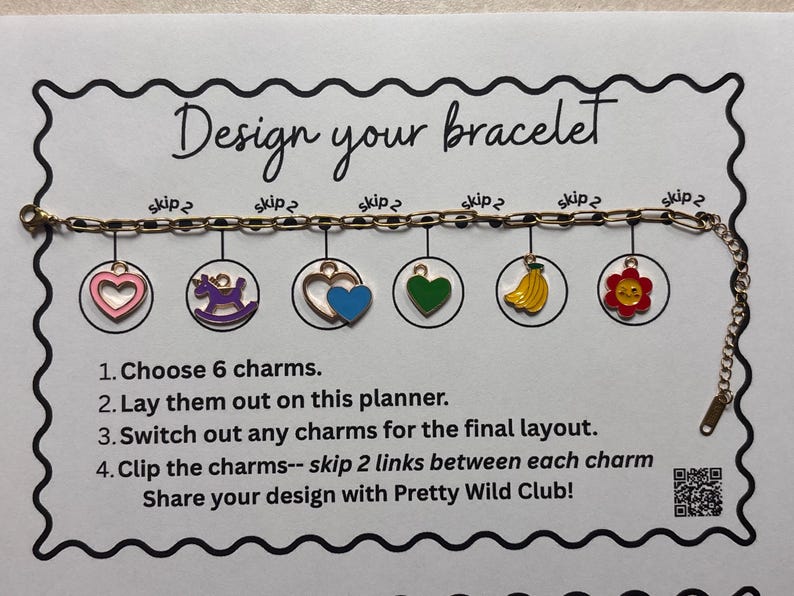 May include: A gold-toned metal chain bracelet with six colorful enamel charms including a pink heart, purple unicorn, blue heart, green heart, yellow bananas, and a red flower. The bracelet is displayed on a white card titled Design your bracelet.