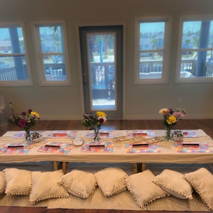 May include: A low-profile dining table setup with a beige tablecloth, featuring colorful patterned placemats, small vases of mixed flowers, and silver disco balls. Several beige corduroy floor cushions with pom-pom trim are arranged around the table.