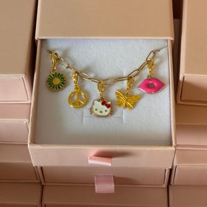 May include: A gold-toned chain bracelet featuring five dangling charms: a green and yellow daisy, a gold peace sign, a white Hello Kitty face with a red bow, a gold butterfly, and a bright pink pair of lips, all presented inside a light pink box.